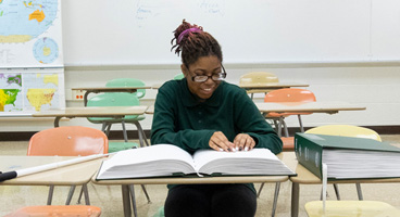 A teenage girl smiles as she reads from a Braille book in a classroom.